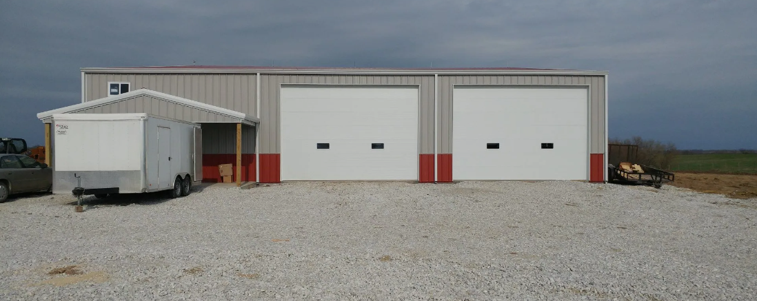 A large gray industrial building with two garage doors and a small trailer parked in front on a gravel lot under a cloudy sky.