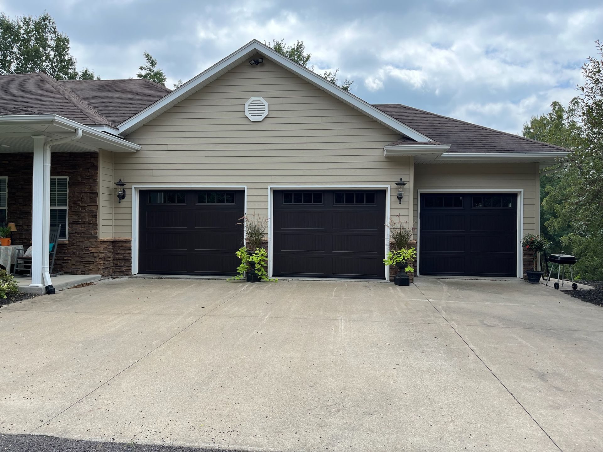 Three-car garage with black doors and light beige siding. Concrete driveway. Cloudy sky.