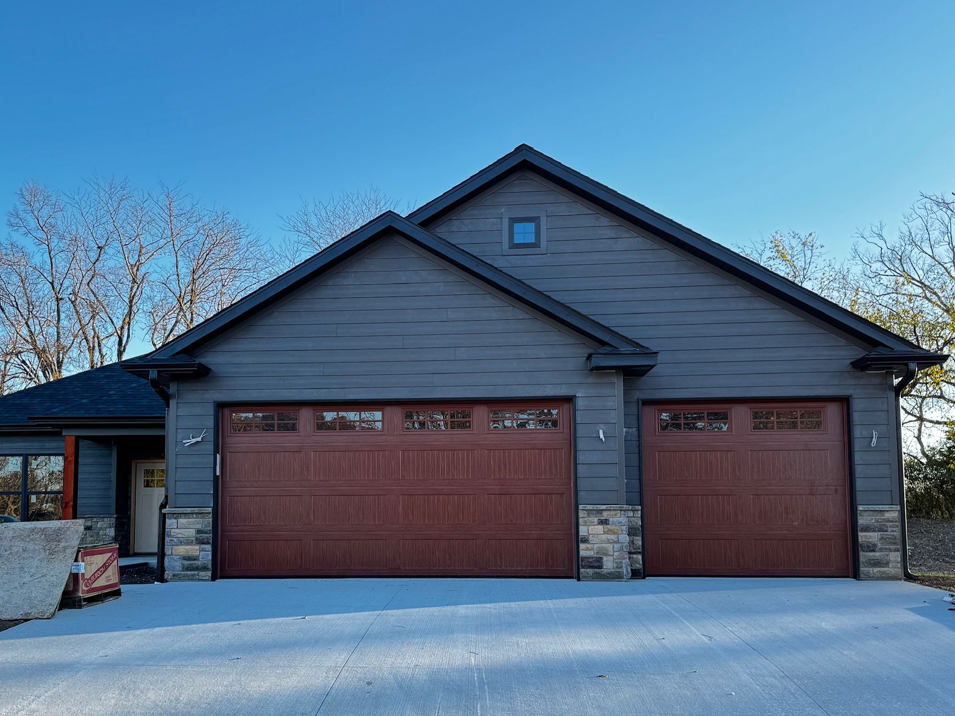 Two-car garage with maroon doors and gray siding, stone accents, on a clear day.