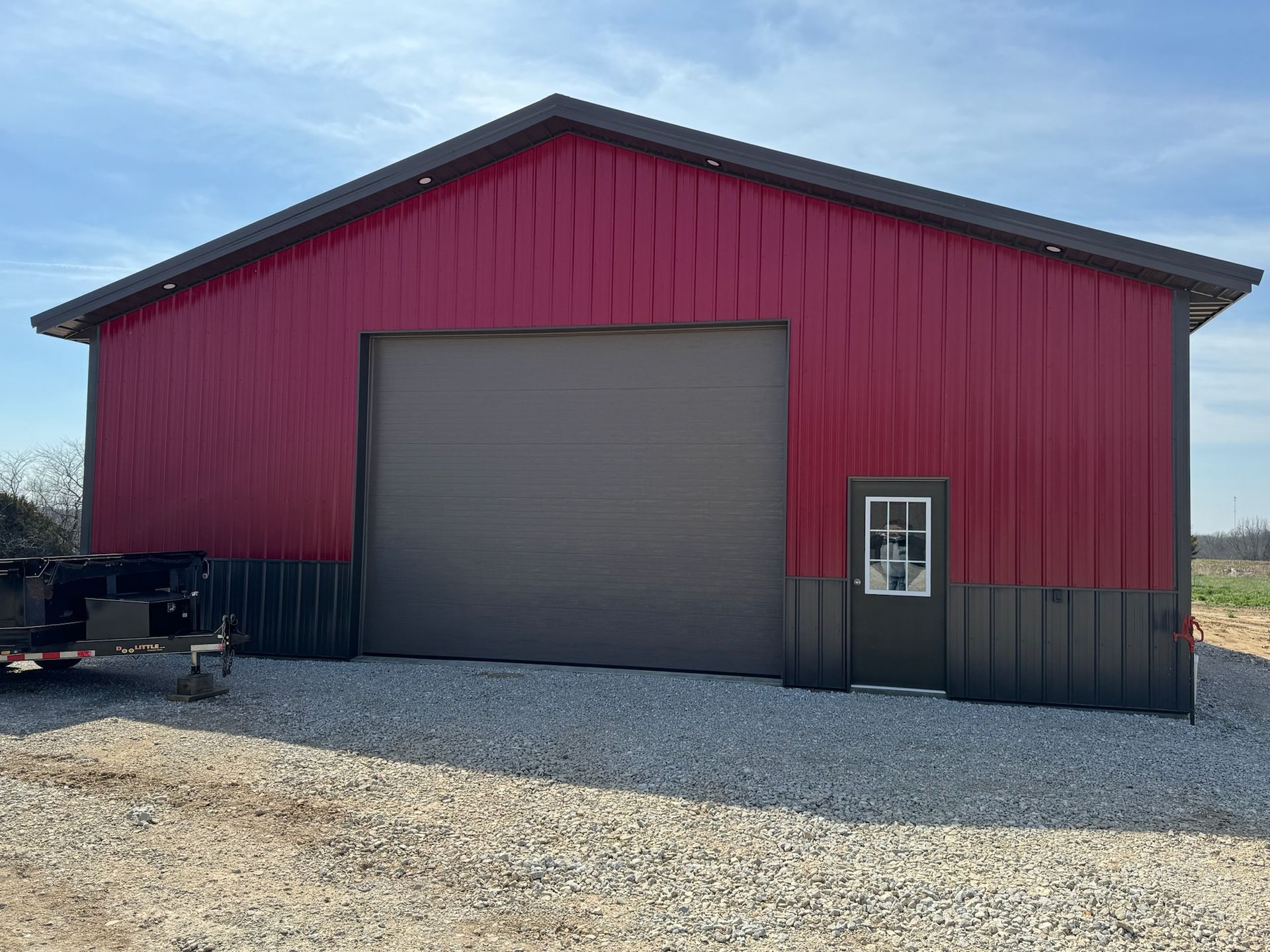 Red and gray metal building with a large garage door, a small door, and gravel ground.