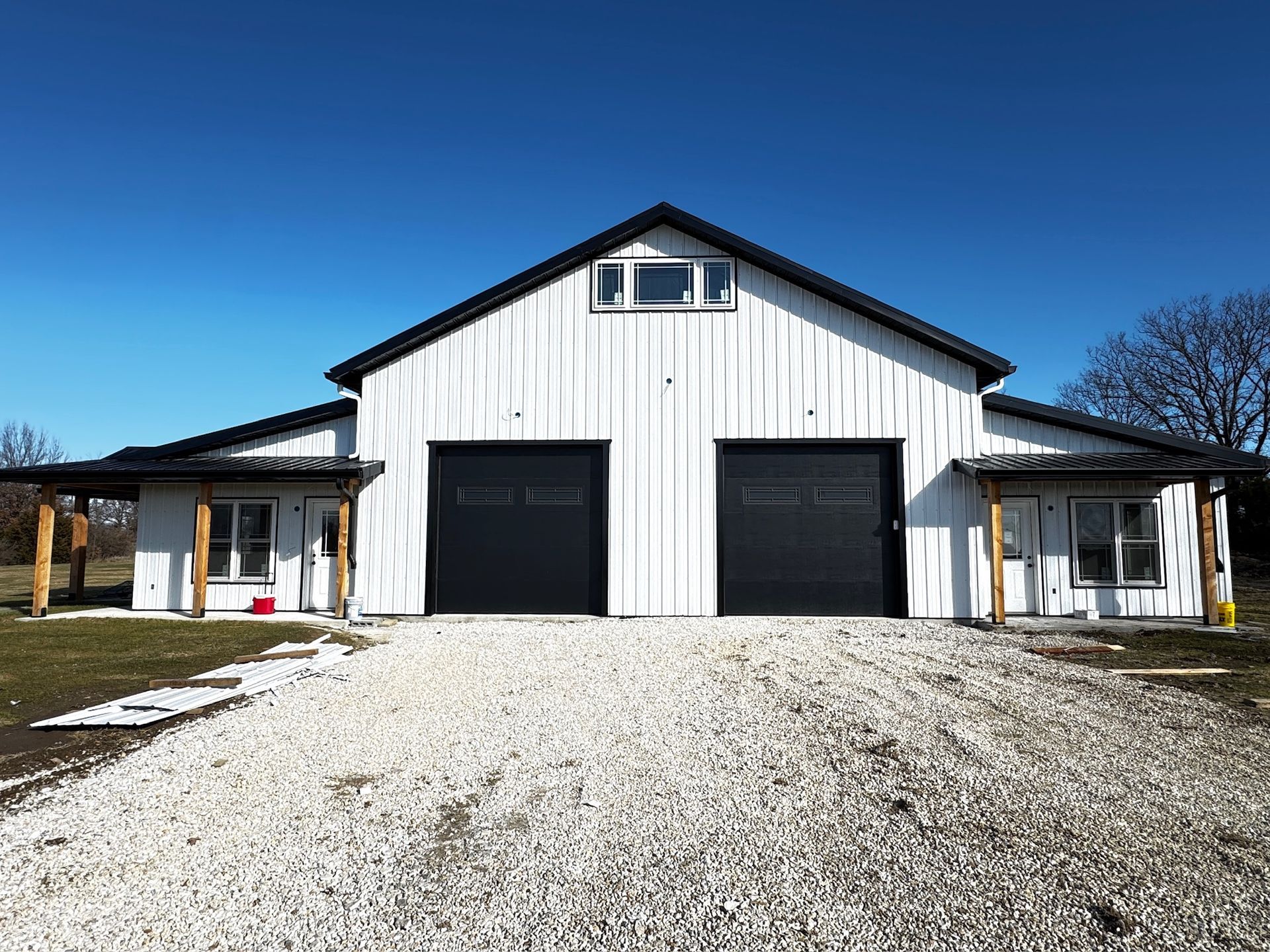 White barn-style building with two dark garage doors and a gravel driveway under a blue sky.