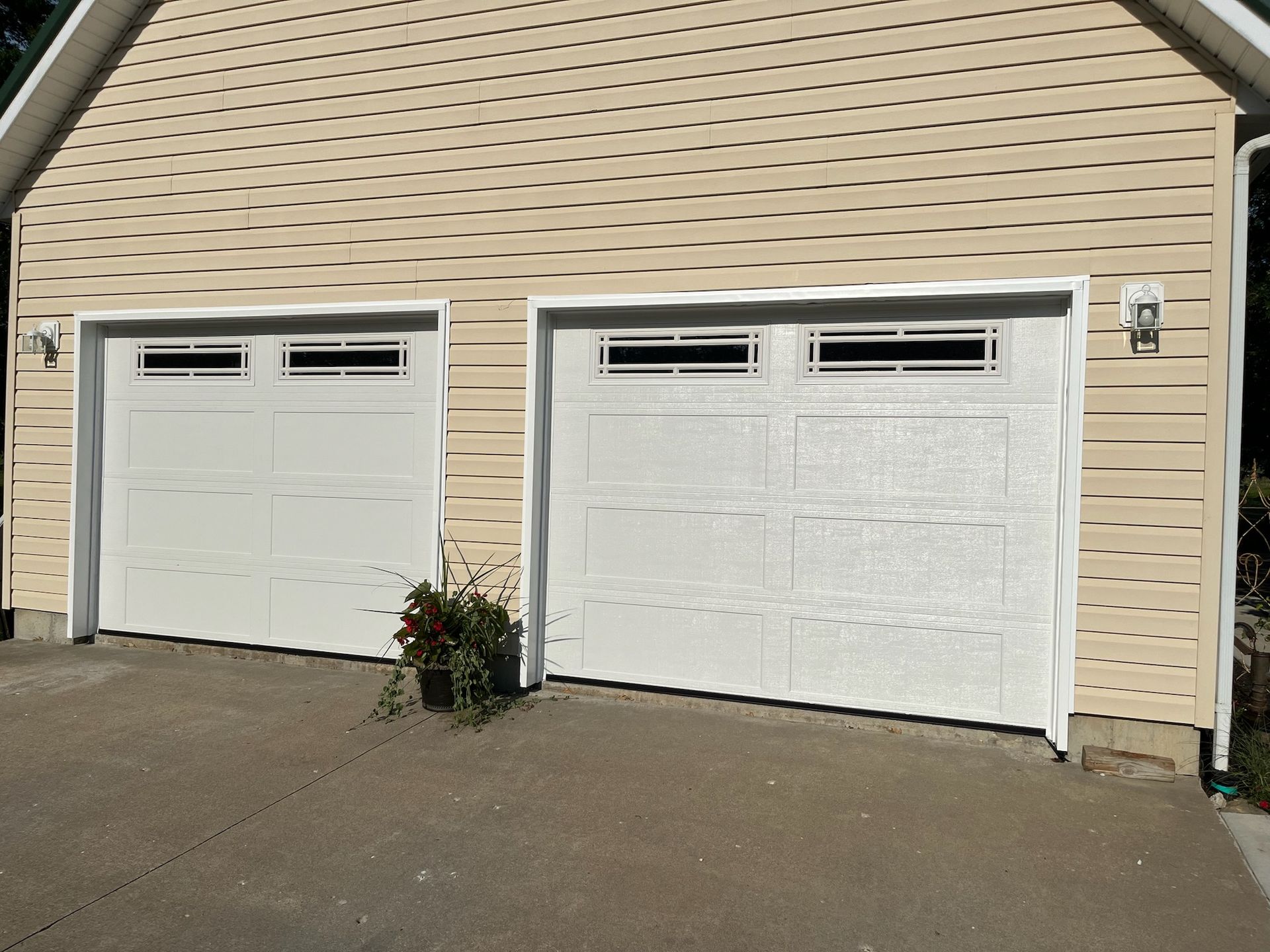 Two white garage doors on a tan house with a concrete driveway, a flower pot, and a light fixture.