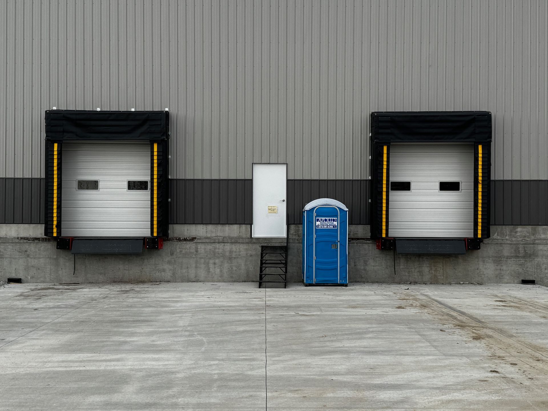 Exterior view of a warehouse with loading docks, a white door, a blue portable toilet, and a small set of stairs.