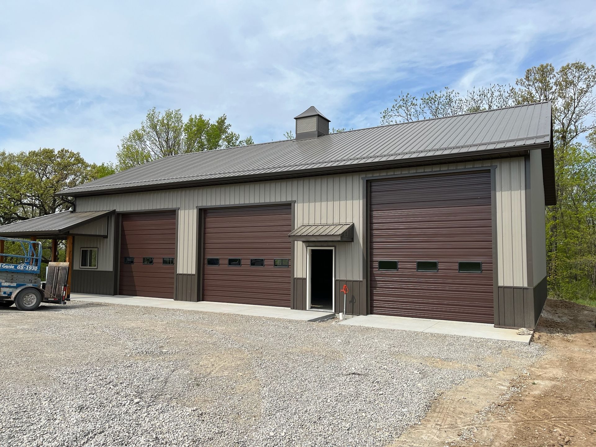 Three-bay metal garage with brown doors, tan siding, and a gray roof, set on gravel.