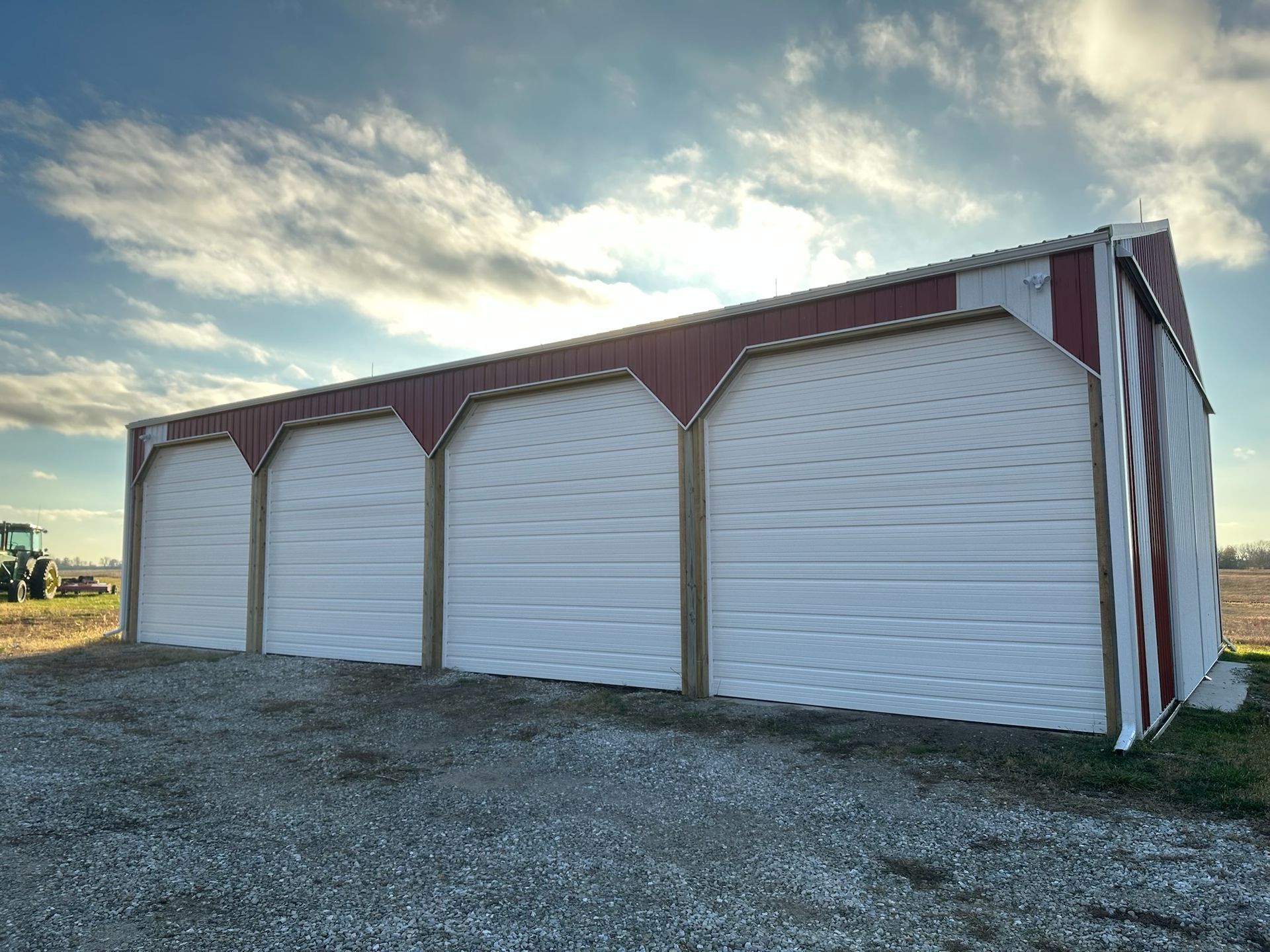Large white and red metal garage with four doors, set on a gravel lot under a cloudy sky.