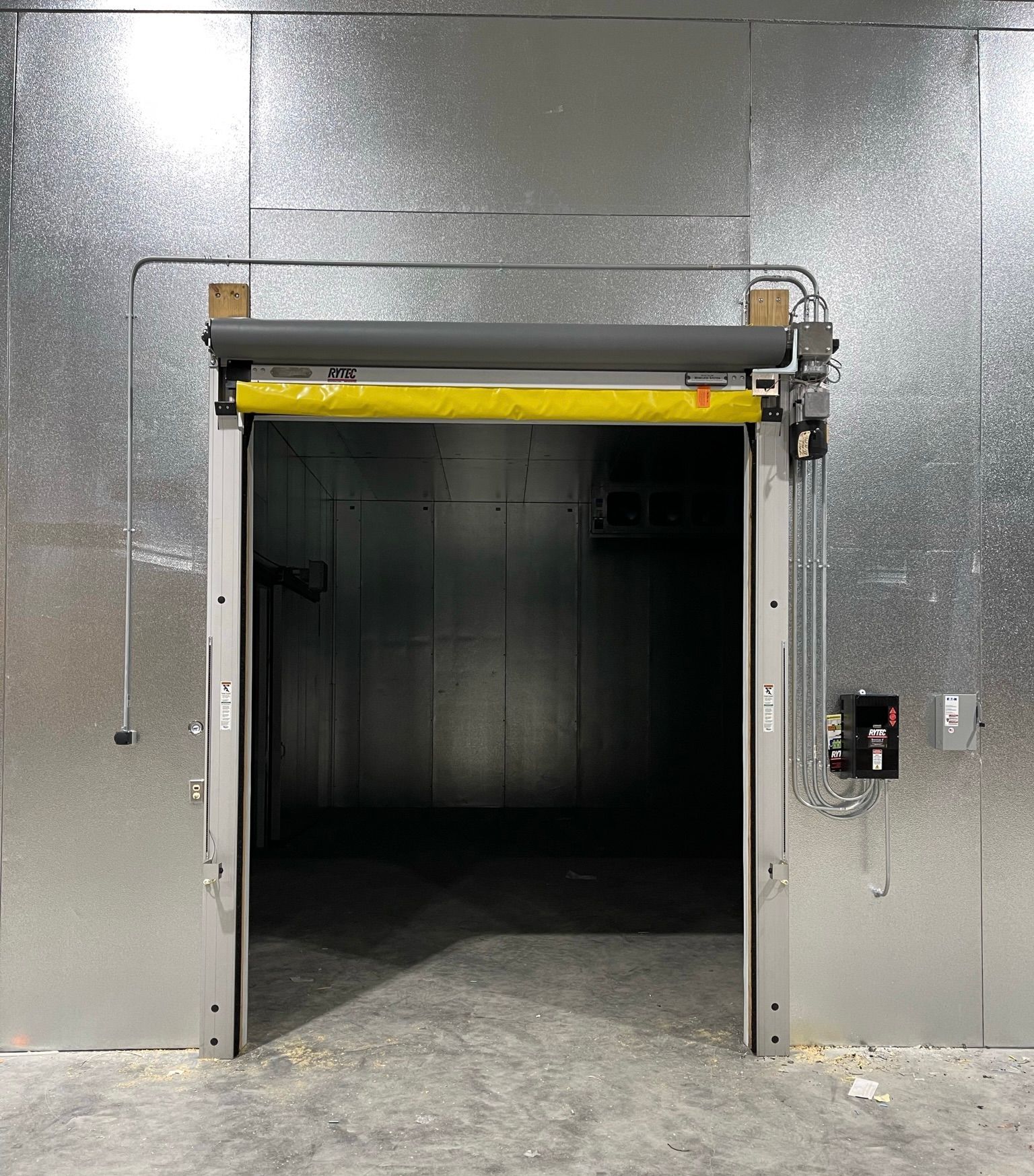 Metal walk-in cooler doorway open, revealing dark interior. Yellow safety bar at top, metal walls.