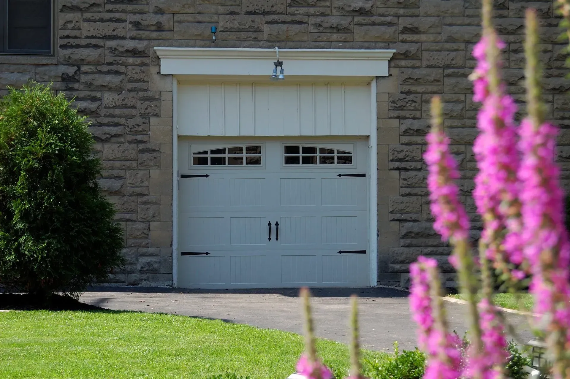 White garage door with decorative trim on a stone building, purple flowers in the foreground.