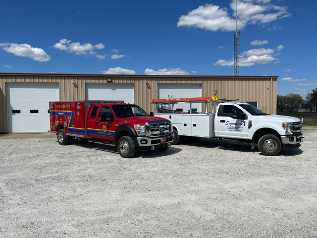 Two trucks, a red ambulance and a white service truck, parked in front of a building with garage doors, under a blue sky.