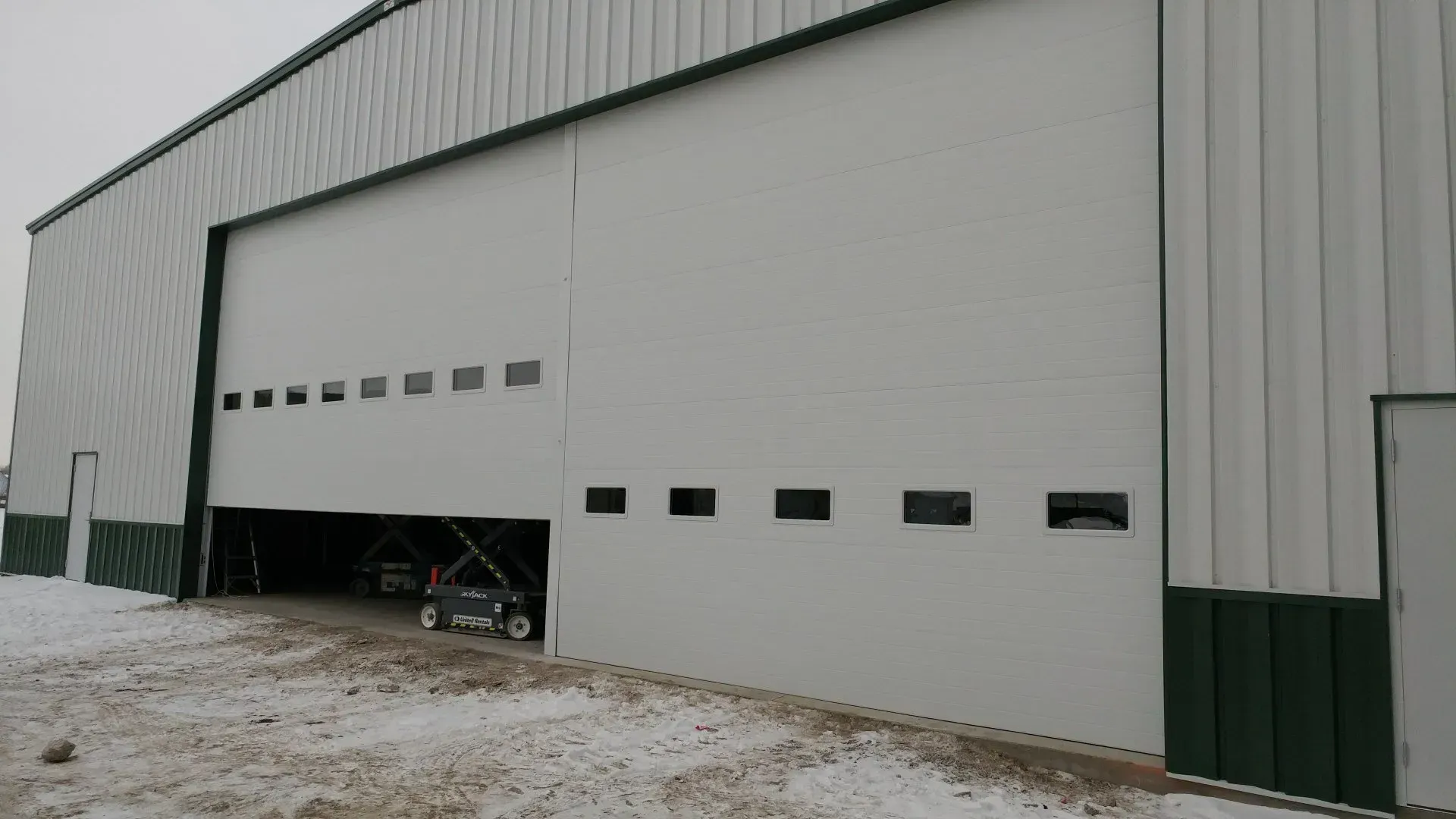 Large white building with open sliding door. A small vehicle is visible inside. Green trim. Snow on ground.