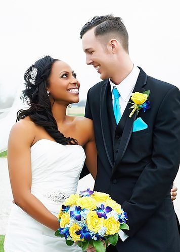 A bride and groom are posing for a picture and the bride is holding a bouquet of yellow and blue flowers.