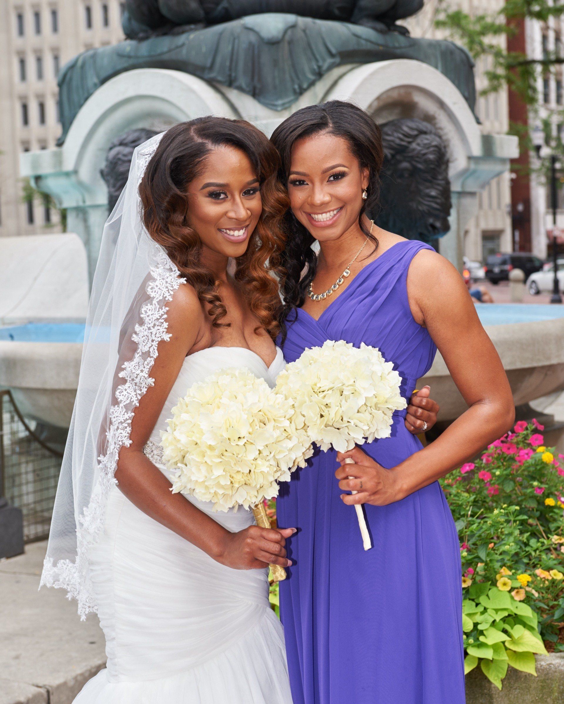 A bride and her bridesmaid pose for a picture in front of a fountain