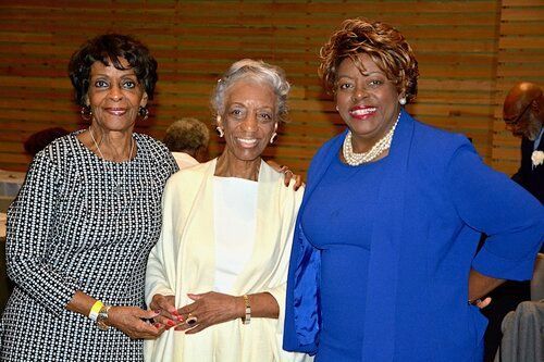 Three women smiling for the camera in a room; one in blue, one in white, one in black and white