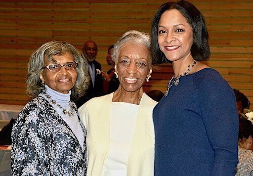 Three women smiling together in a room with light wood paneling in the background