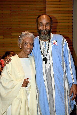 Woman in cream dress with man in blue tunic, both smiling, arm around each other