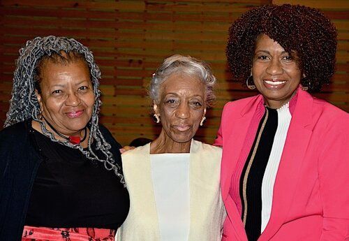 Three smiling women pose close together. One wears a pink blazer