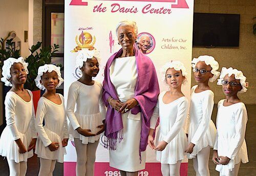 Woman in white with dancers in white dresses and bonnets in front of a banner