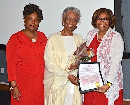 Three women in red and cream attire, one holding a certificate and flowers