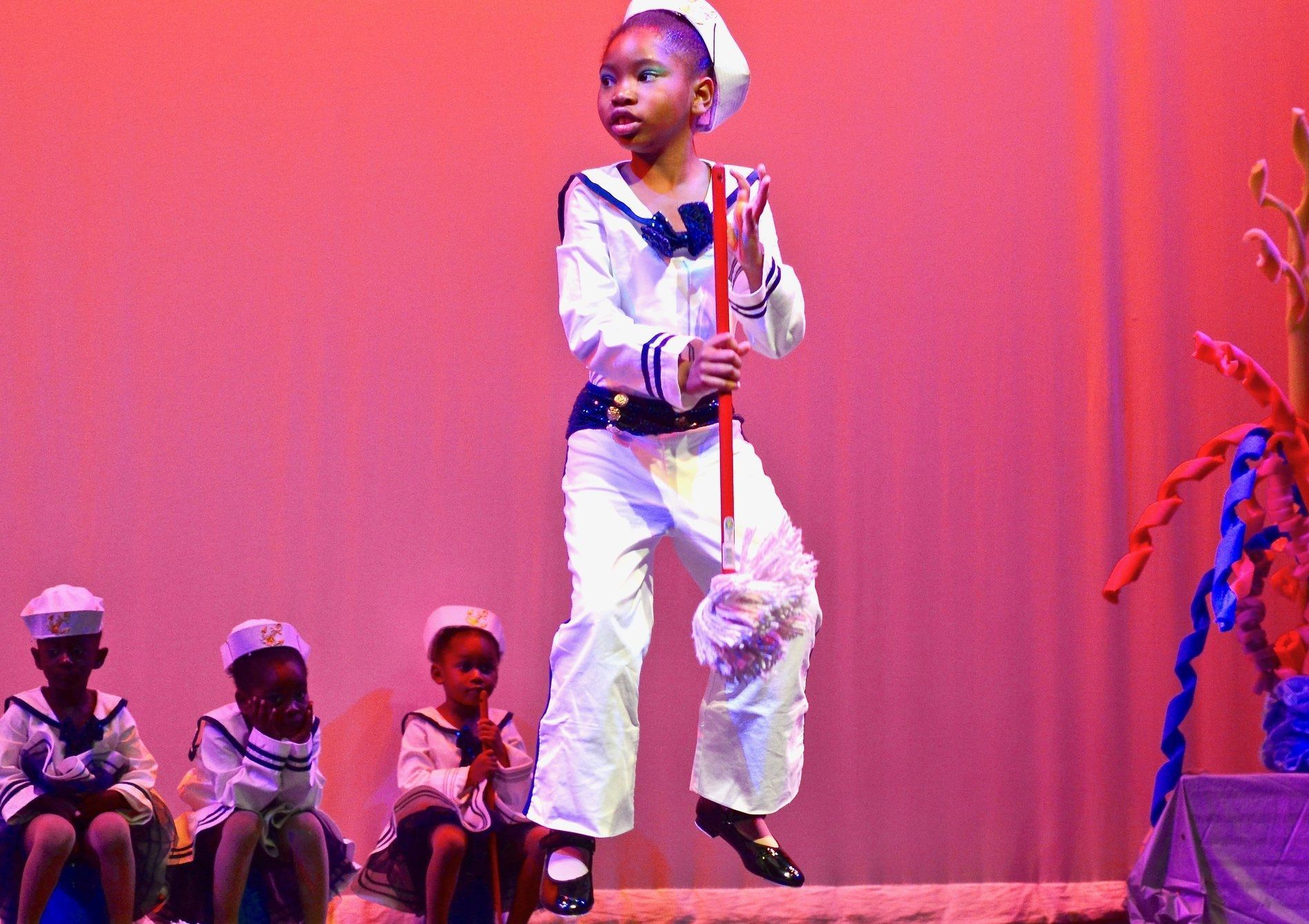 A child in a sailor costume dances on stage with a red staff