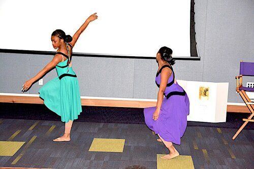 Two dancers in colorful dresses performon a stage with a screen, one arm raised.