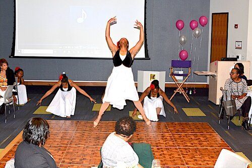 Dancers in white dresses and black vests perform on a wooden dance floor