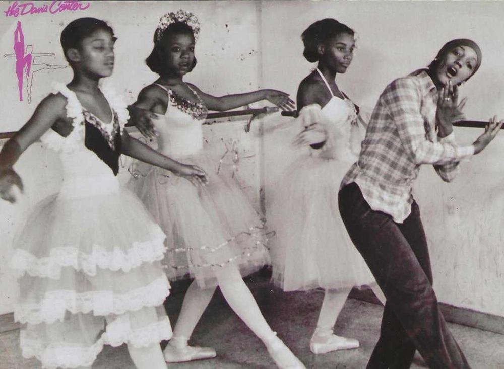 Four ballet dancers at a barre; three in tutus, one in casual wear, practicing dance.