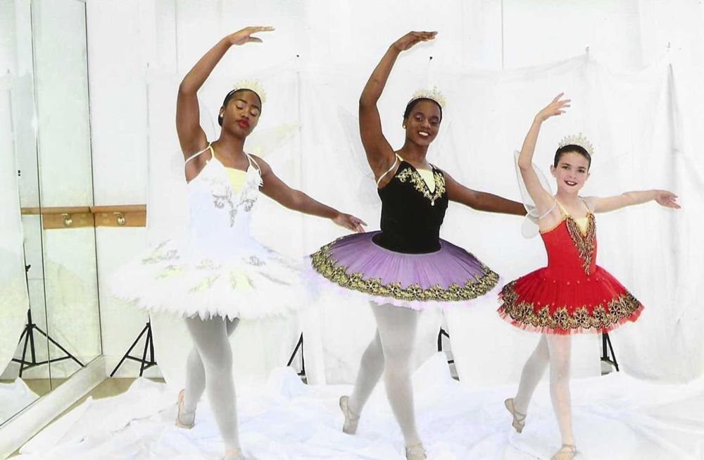 Three ballerinas in tutus and tiaras, posed in a studio with raised arms.