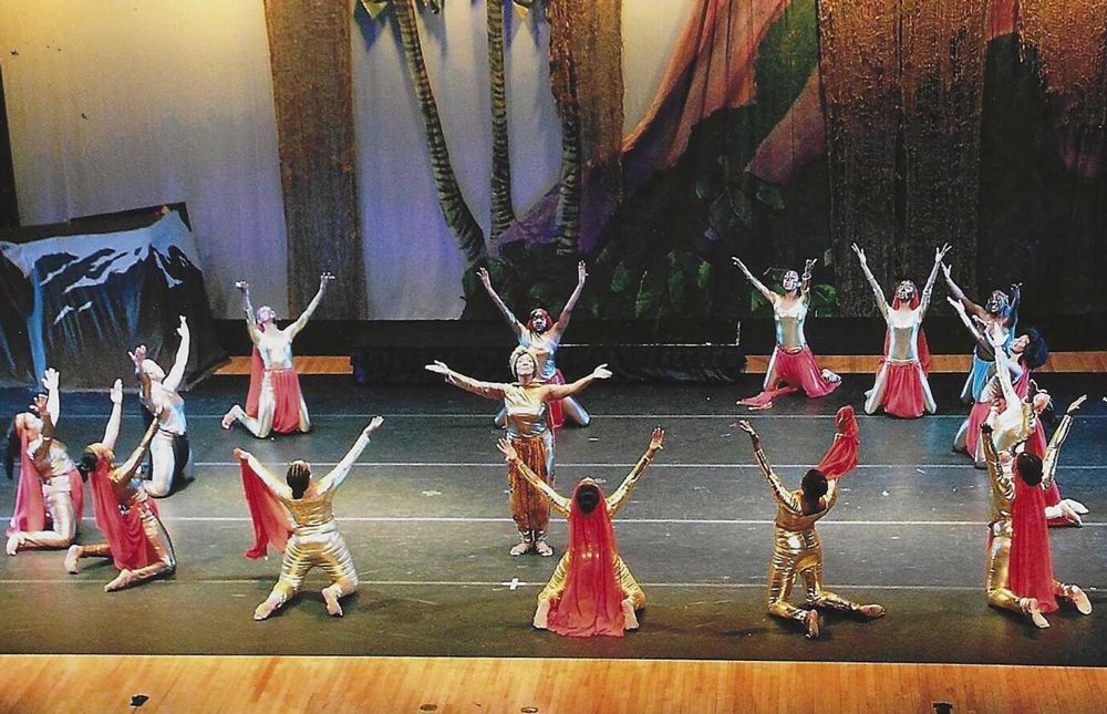 Dancers on stage with arms raised, wearing colorful costumes, performing in front of a painted backdrop.