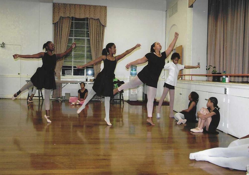 Ballet dancers in a studio, performing arabesque. Wooden floor, large window, dancers in black dresses and tights.