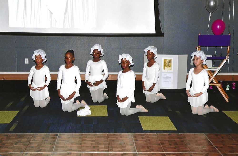 Seven children in white costumes kneel, hands clasped, on a stage. They wear head coverings and tights.