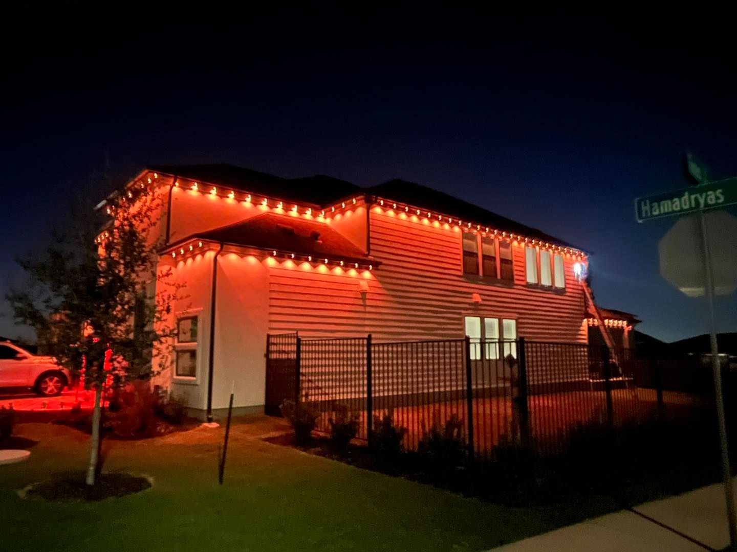 Two-story house with red Christmas lights along the roof at night. Street sign in the corner.