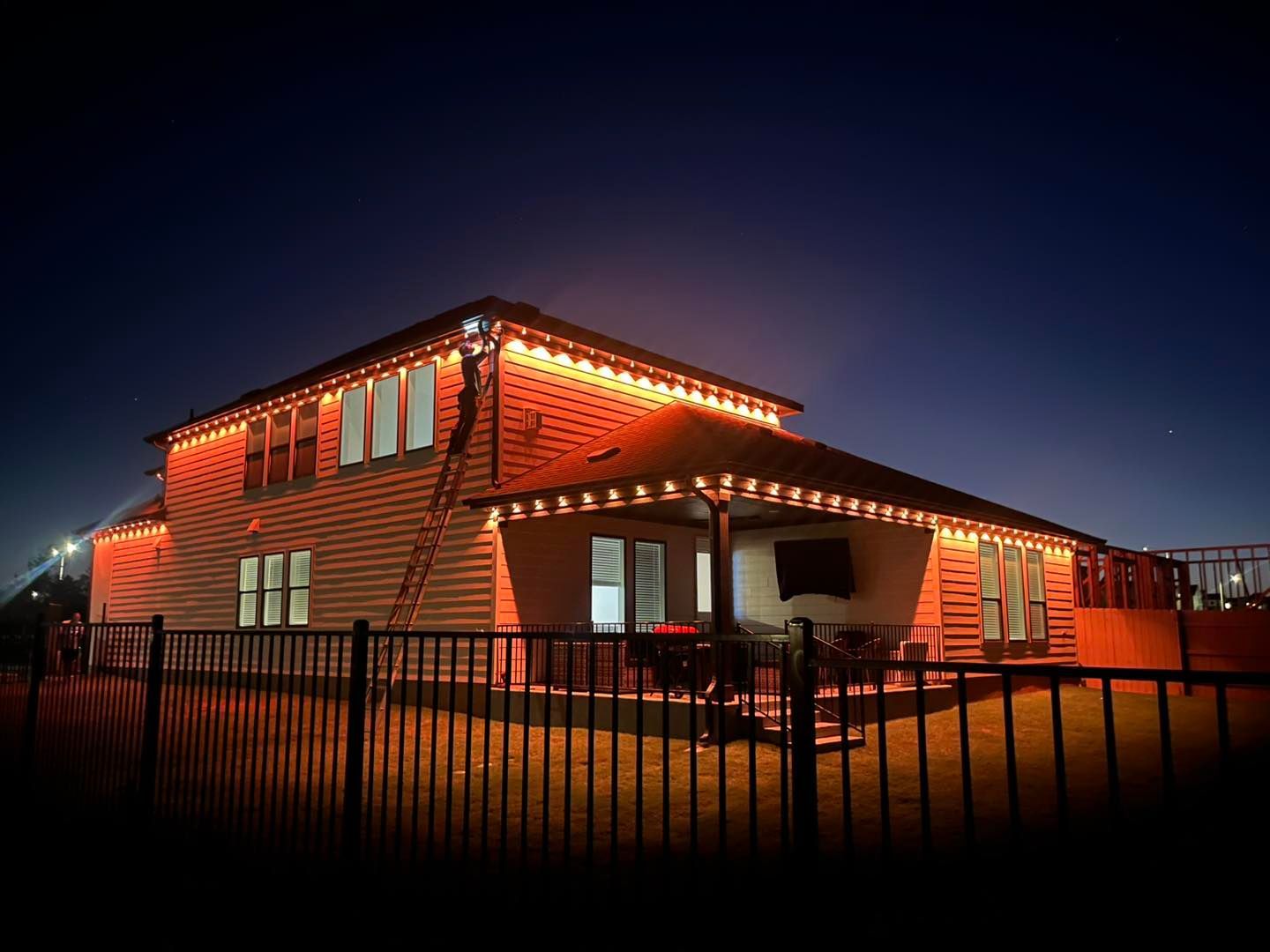 House at night with orange Christmas lights along roof and porch, behind a black fence.