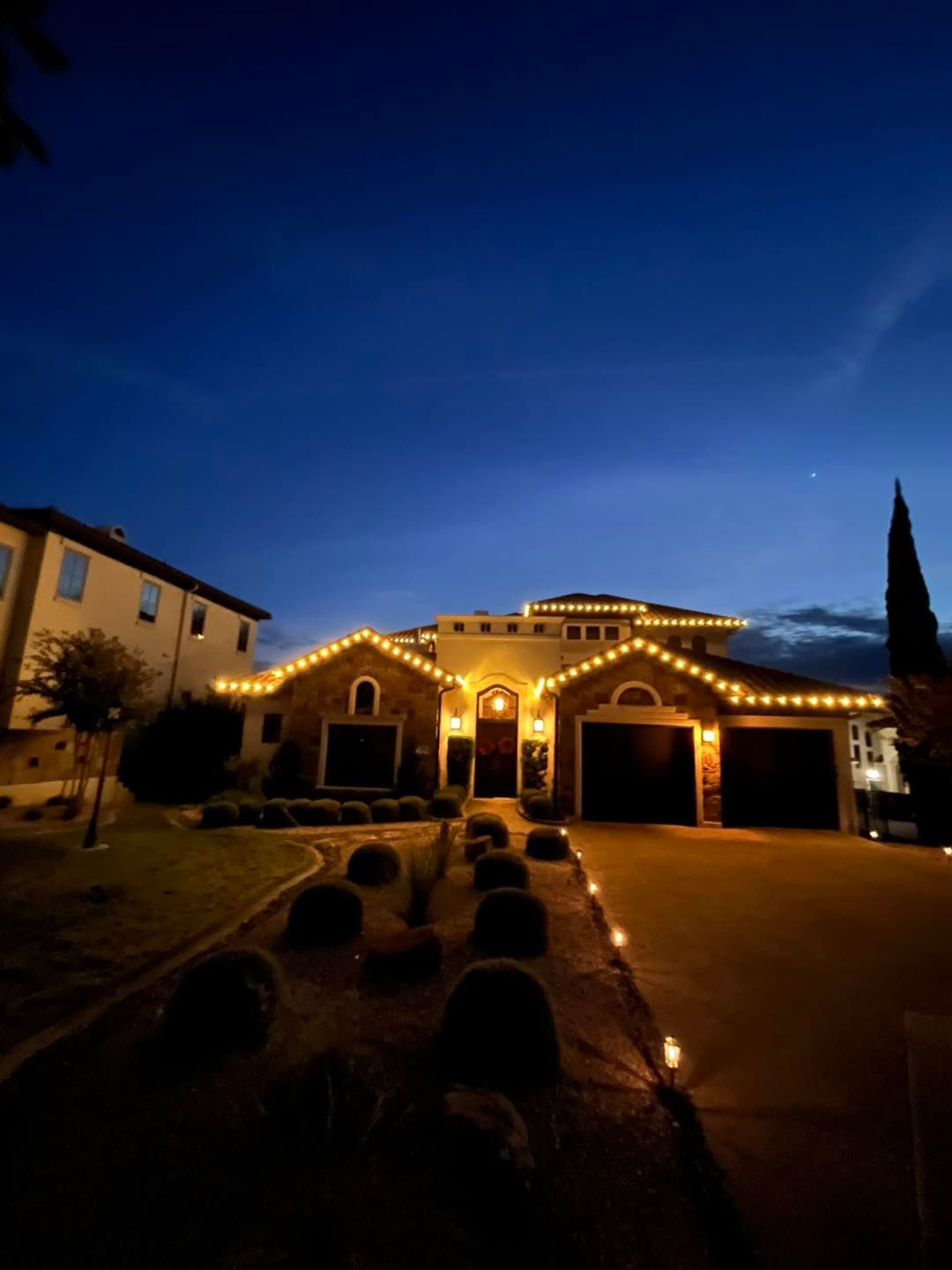 House at night with gold lights outlining the roof and pathway. Dark blue sky.