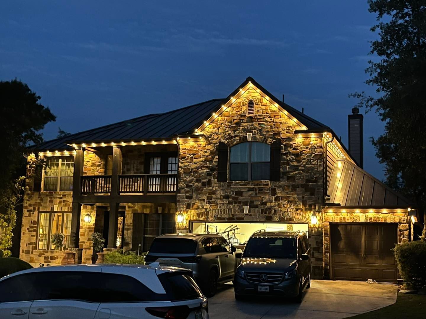 House decorated with string lights at dusk; vehicles parked in front.