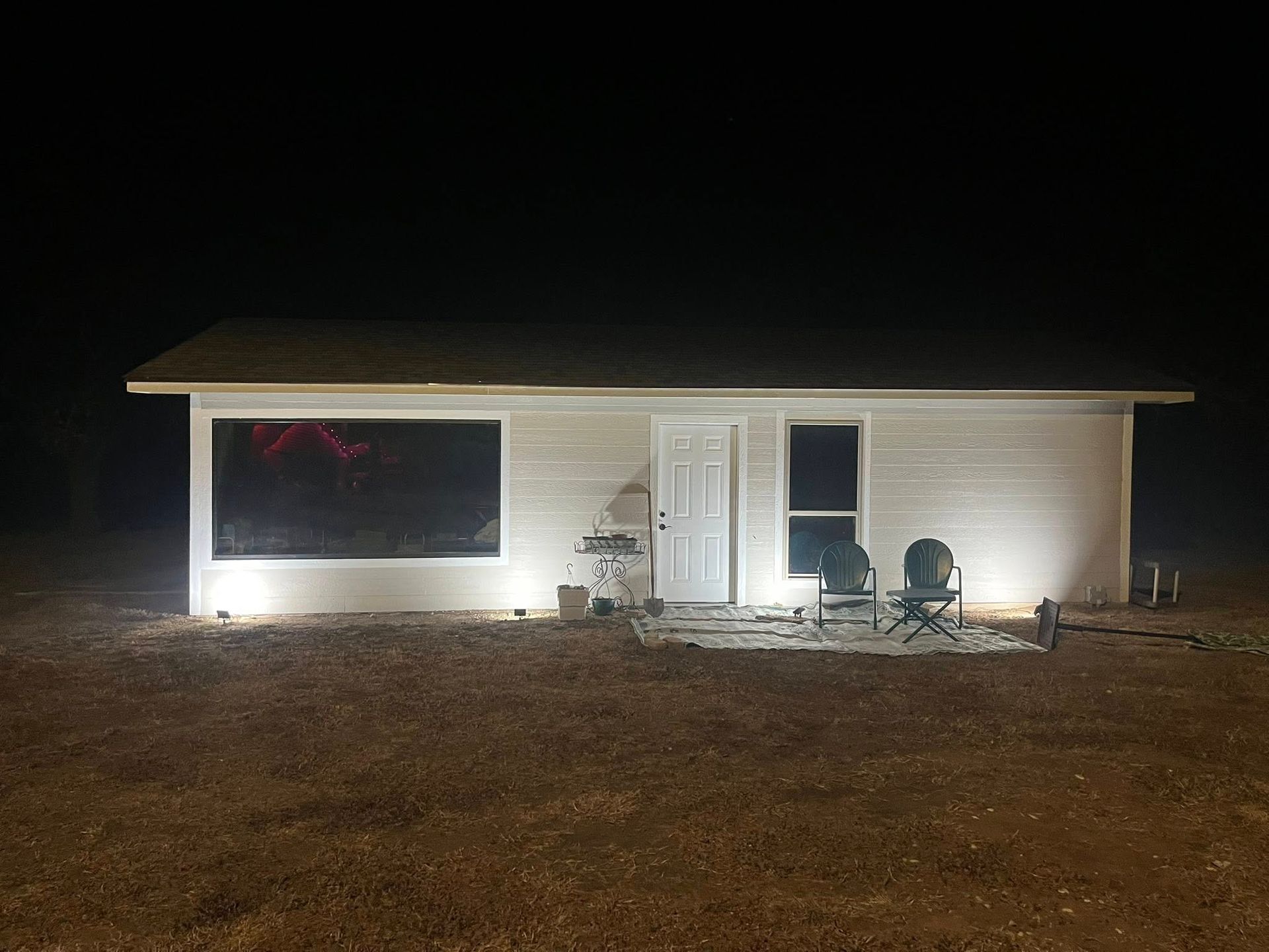 White building with lights at night. A large window and a door are visible. Two chairs sit in front.