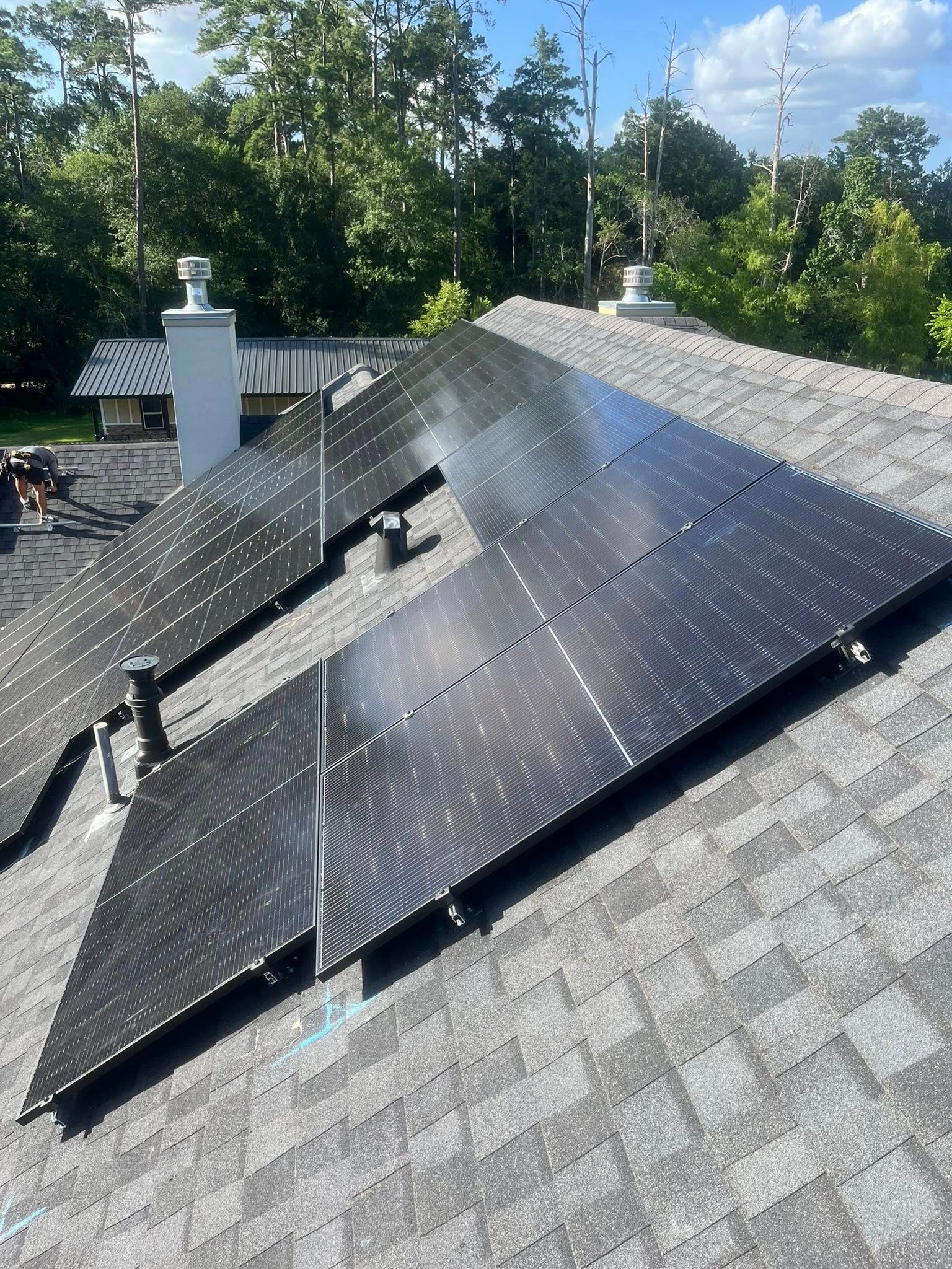 Solar panels installed on a gray shingle roof against a background of trees and blue sky.