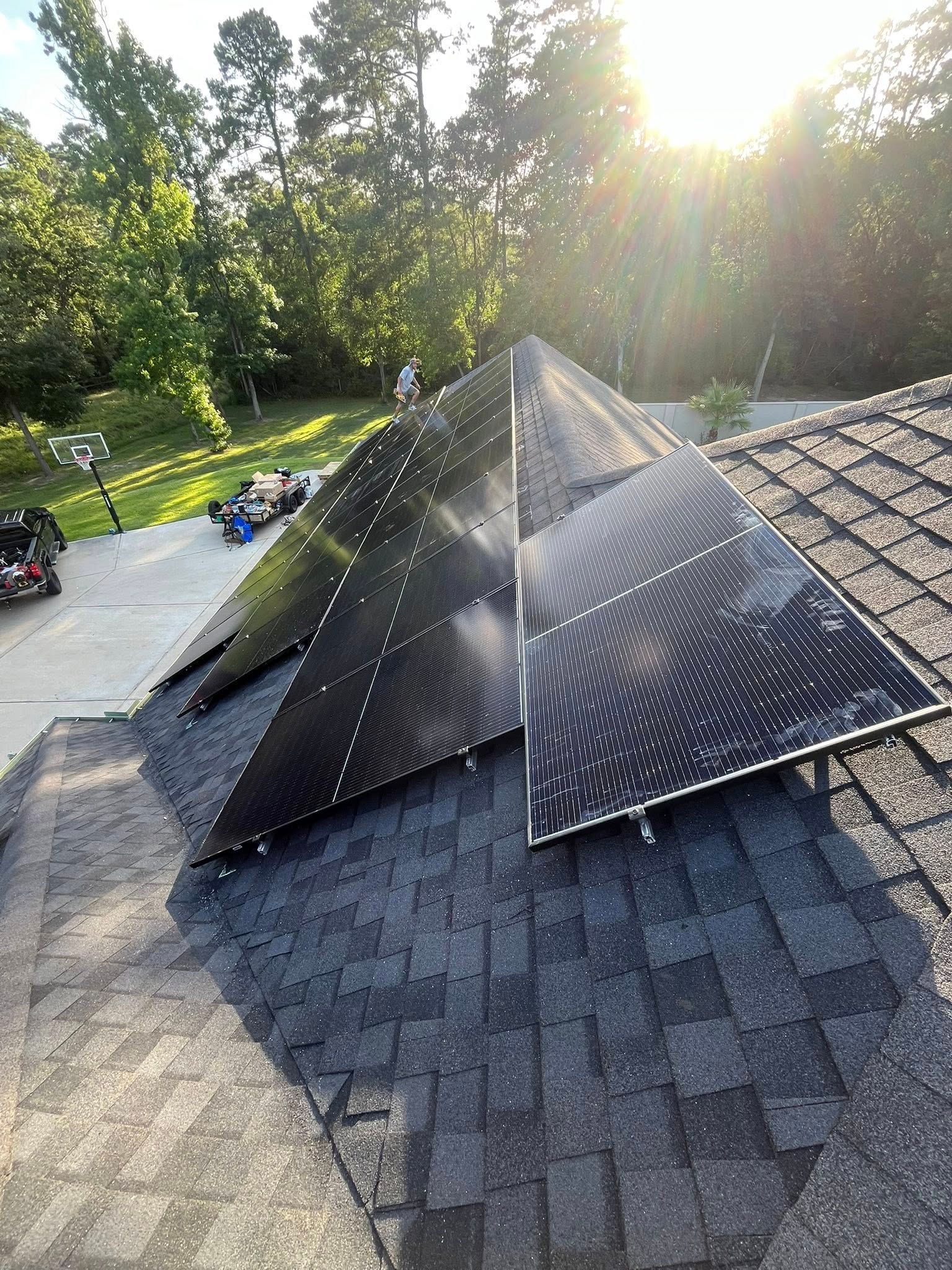 Solar panels installed on a dark shingled roof, trees and vehicles in the background. Bright sunlight.