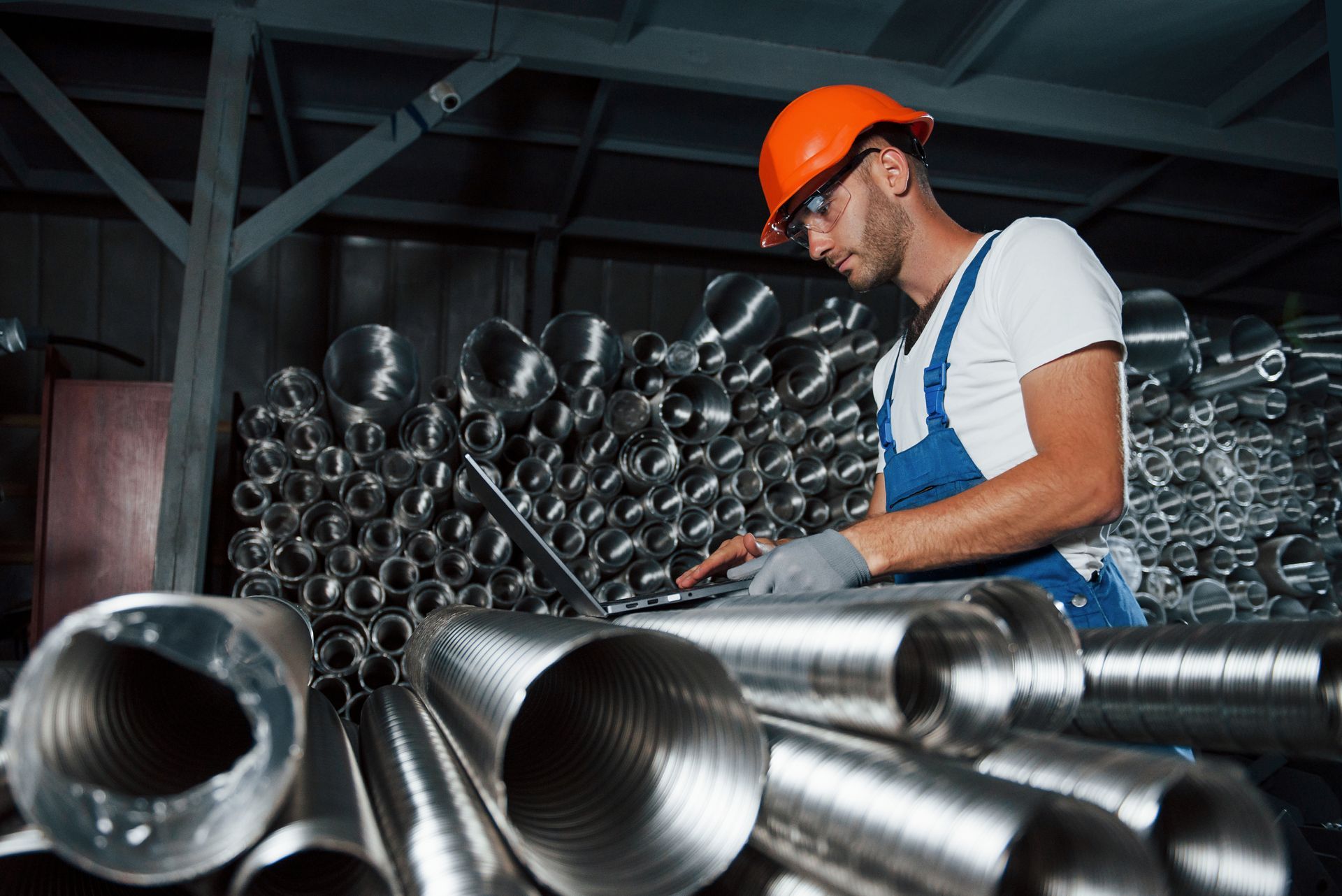 A builder in uniform works on a laptop in a warehouse full of steel pipes.