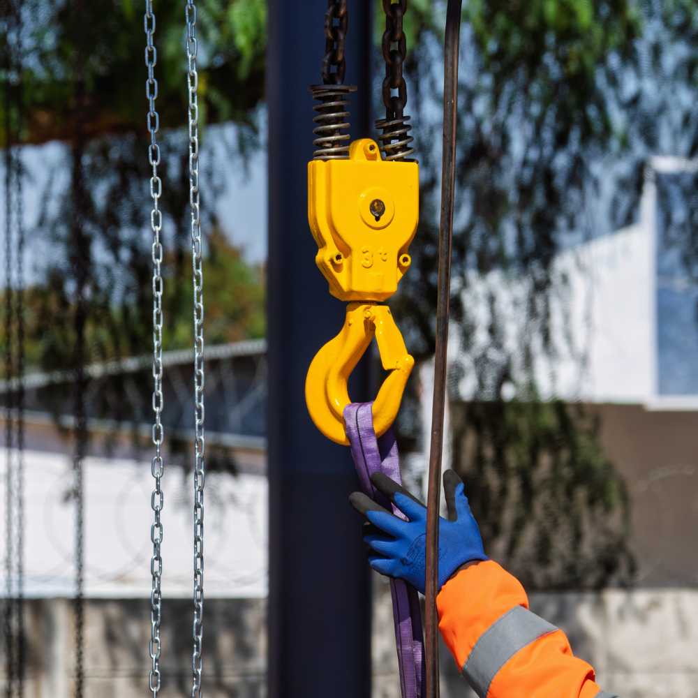 Yellow crane hook lifting a purple strap, with gloved hands guiding it on a worksite
