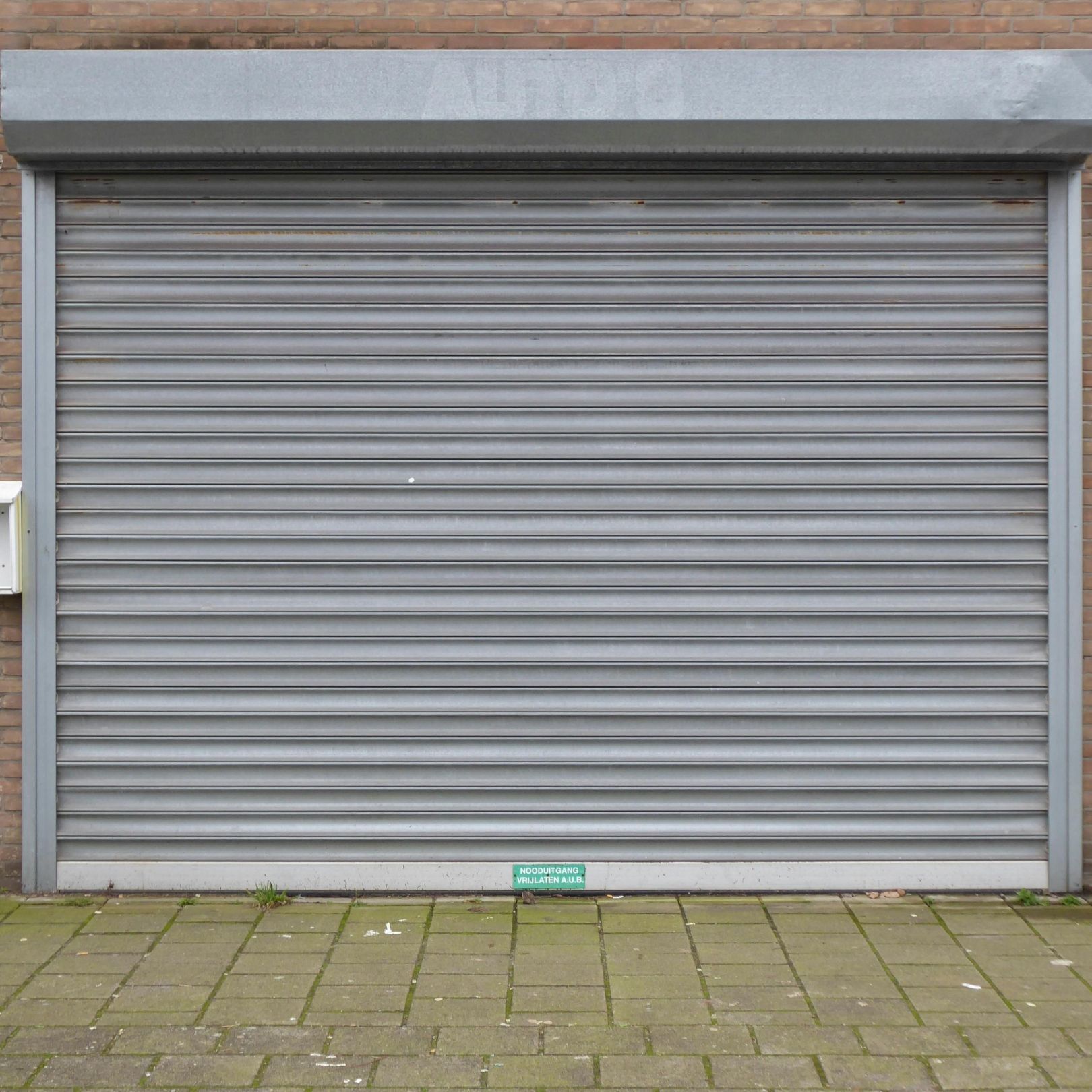 Closed gray metal roll-up garage door in a brick wall with a tiled pavement in front