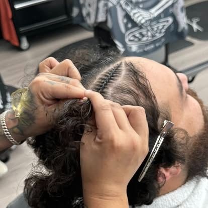 Hands braiding hair on a person's head, close up. The hair is parted, with one side styled into cornrows.