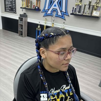 Woman with braided hair, wearing glasses and a black shirt, indoors, in front of a wall with trophies.