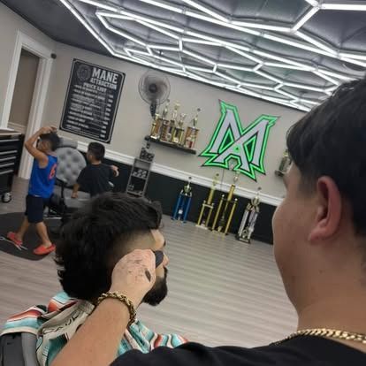 Barber giving a haircut in a barbershop with a logo on the wall. Customers wait in the background.