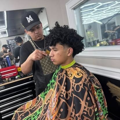 A barber giving a client a haircut in a shop. The client wears a patterned cape.