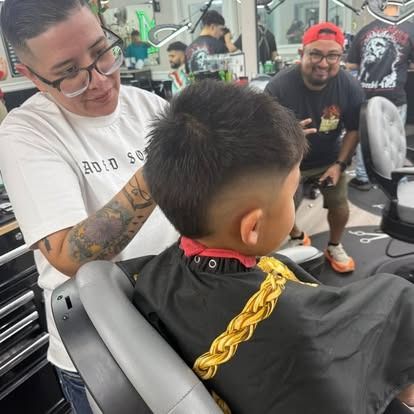 Barber giving a young person a haircut in a shop; client wearing cape.