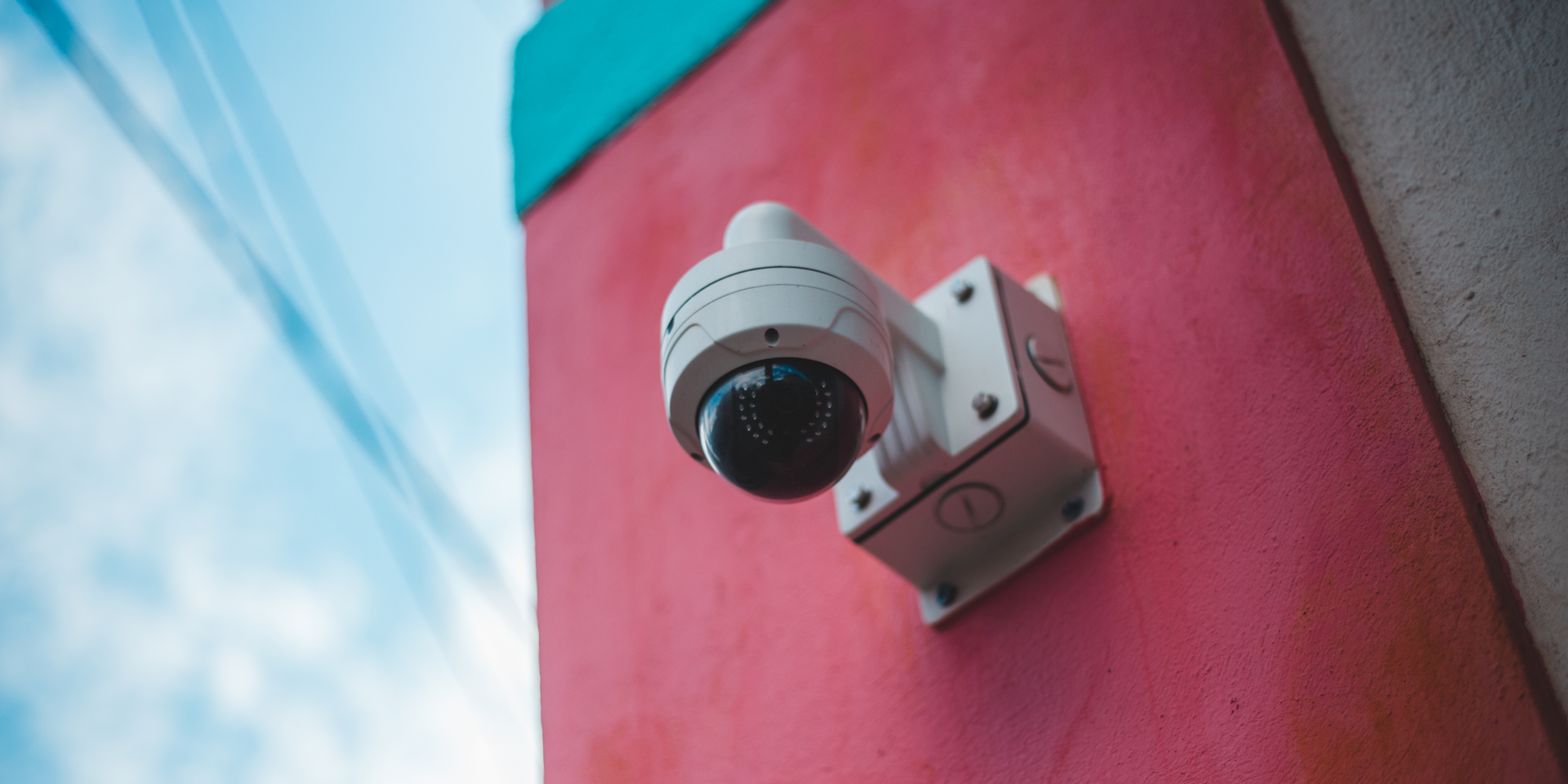 Security camera mounted on a pink wall, with a blue sky in the background.