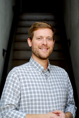 Man in a button-down shirt smiles, leaning against a wooden post. Floral arrangement in background.