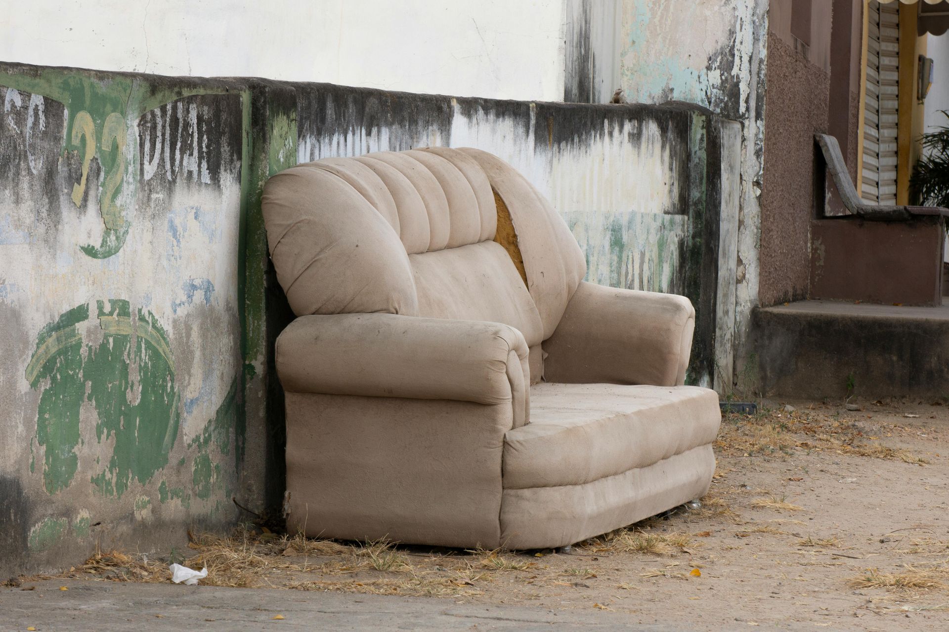 Beige armchair abandoned outdoors beside a weathered wall and steps on a dusty ground