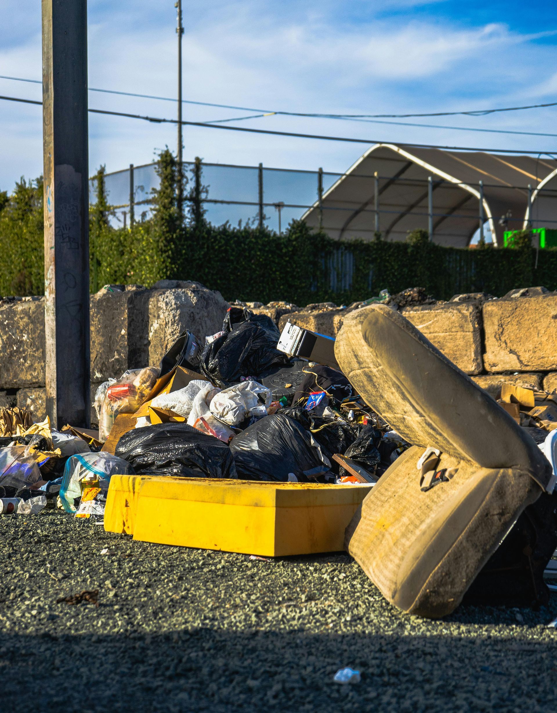 Overflowing trash bags and debris piled beside a yellow dumpster on gravel near a stone wall