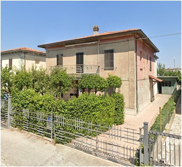 Casa beige a due piani con tetto di tegole rosse, balcone e giardino anteriore dietro una recinzione metallica in una giornata di sole.