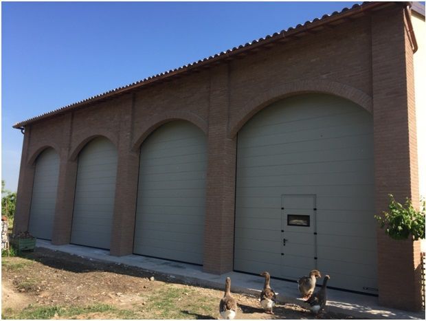 Edificio con quattro porte ad arco e portoni del garage chiusi; oche di fronte.